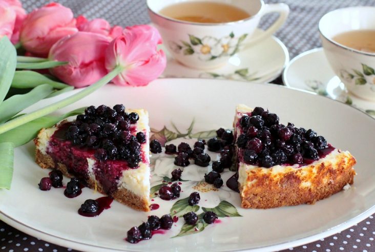 Two slices of lemon cheesecake topped with juicy purple Saskatoon Berry Sauce surrounded by cups of tea and pink tulips.