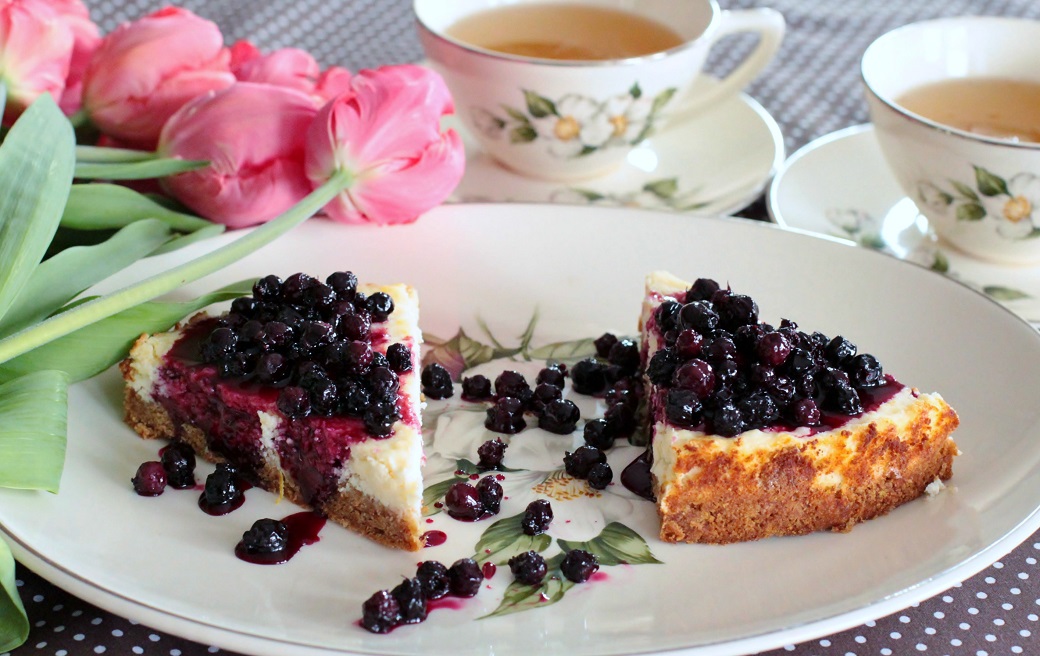 Two slices of lemon cheesecake topped with juicy purple surrounded by cups of tea and pink tulips. Saskatoon Berry Sauce.