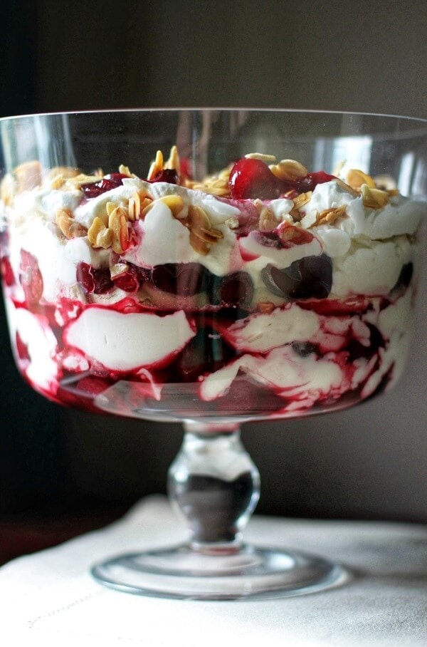 Vertical image of a stemmed trifle bowl filled with red cherries, whipping cream and almonds
