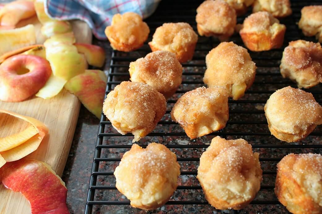 Apple Fritter Bites - Dish 'n' the Kitchen Golden baked apple fritters with chunks of apple sit on a black cooking rack. Apple peelings sit beside.