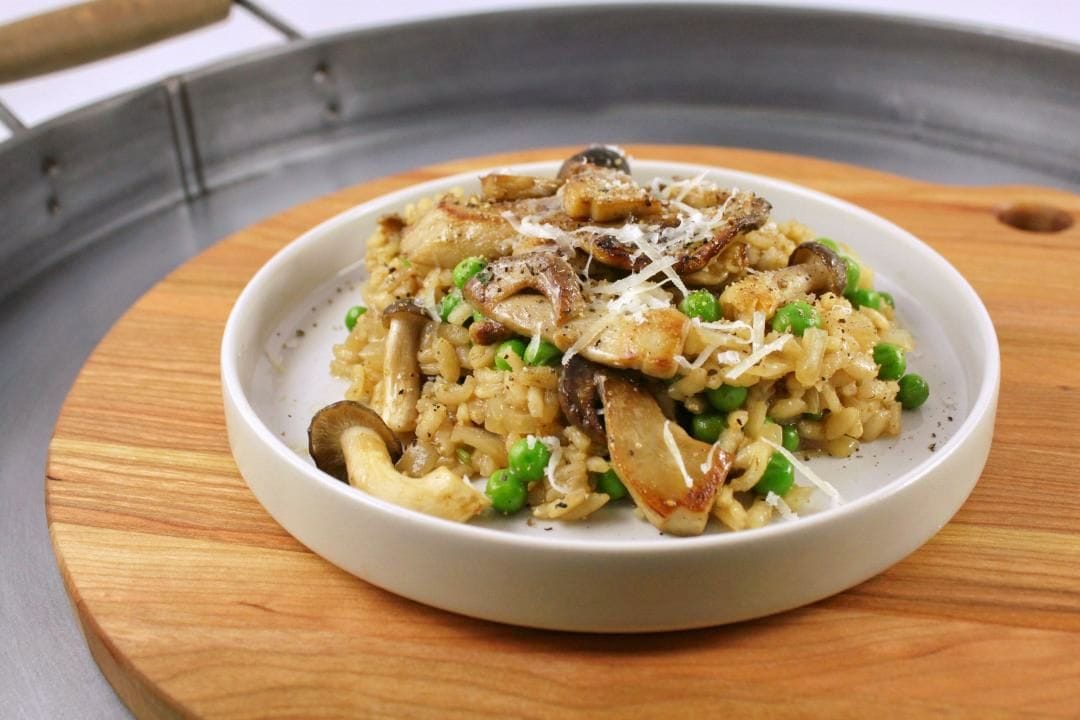 A shallow white bowl filled black garlic risotto, peas, and sauteed mushrooms.