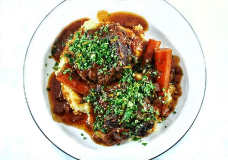 A white plate containing mashed potatoes topped with meaty braised beef short ribs and carrots in a rich gravy topped with a green herb gremolata.