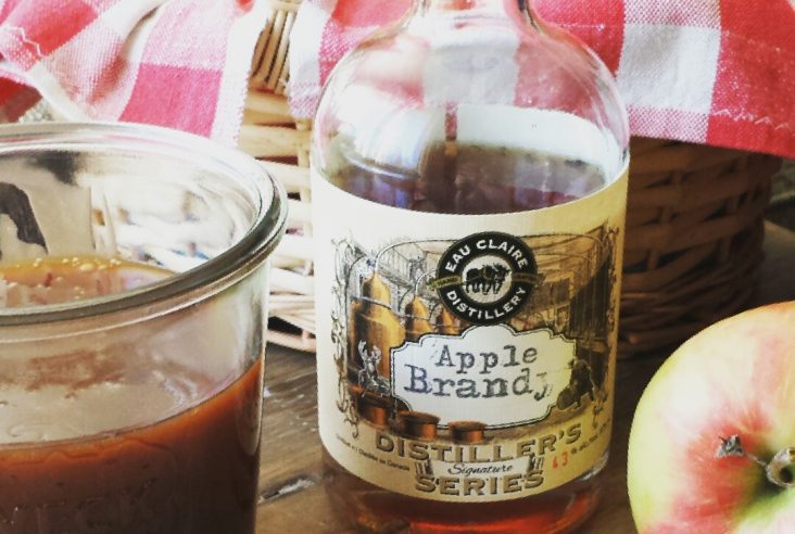 A jar of apple brandy caramel sauce beside apples and a bottle of apple brandy.