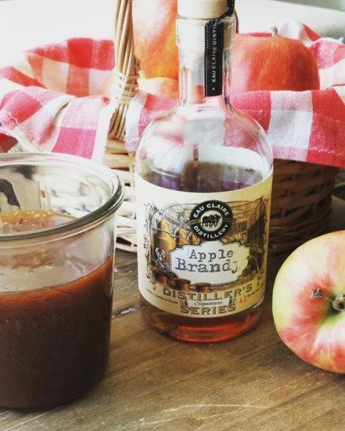 A jar of apple brandy caramel sauce beside apples and a bottle of apple brandy.