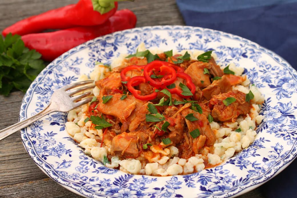 Chicken Paprikash - A delicately flowered china plate containing homemade noodles topped with dark orange chicken paprikash, red peppers and parsley.