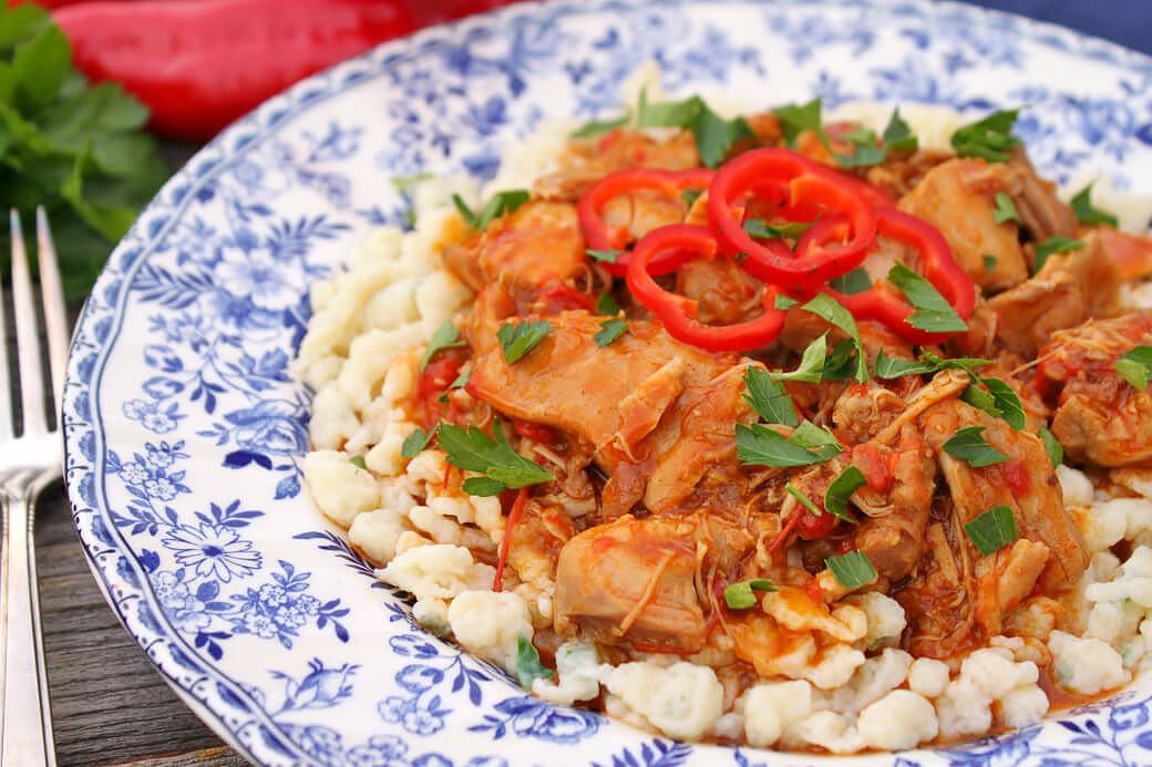 Chicken Paprikash - A delicately flowered china plate containing homemade noodles topped with dark orange chicken paprikash, red peppers and parsley. 