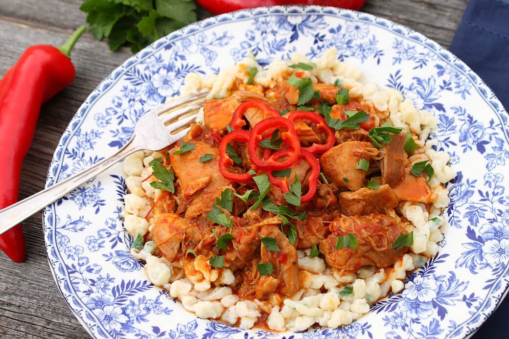 Chicken Paprikash - A delicately flowered china plate containing homemade noodles topped with dark orange chicken paprikash, red peppers and parsley. 
