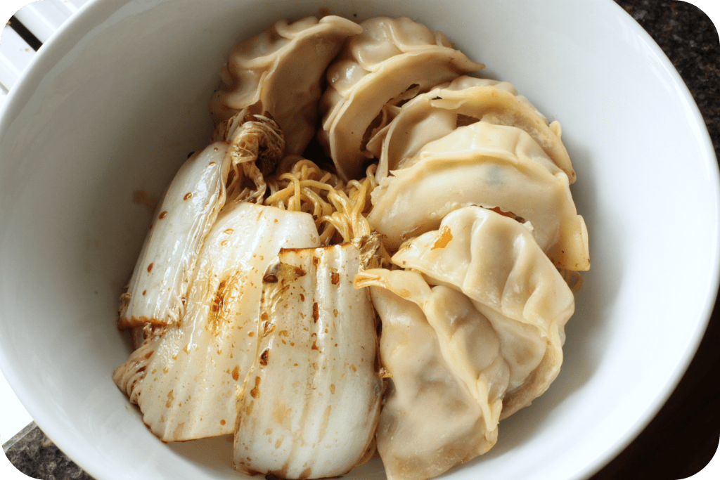 A white bowl containing noodles, cabbage and pan fried pork dumplings.