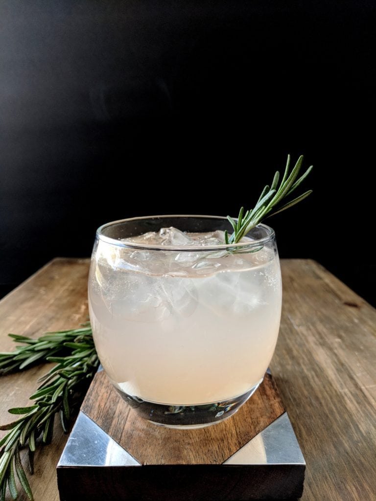 A light pink Mastical Maia cocktails garnished with fresh rosemary and made with Skinos Mastiha, grapefruit, and rosemary syrup
