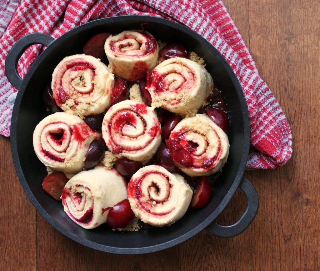 Sourdough Plum Cinnamon Rolls- 8 unbaked rolls with red plums in a black baking pan.