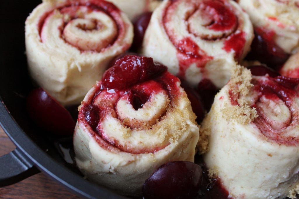 Sourdough Plum Cinnamon Rolls- Close up of unbaked rolls and red plums in a black round pan.
