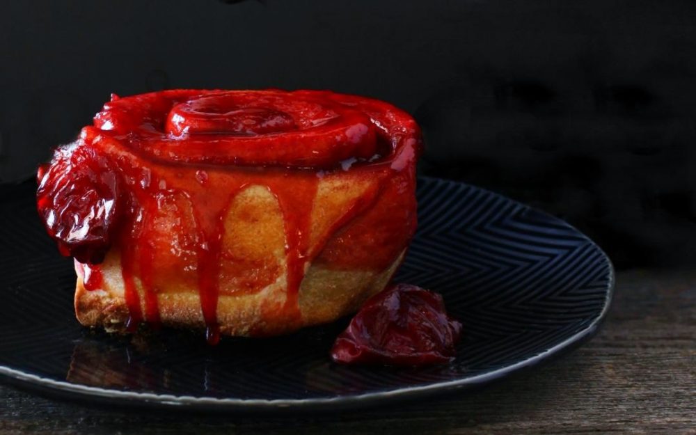 Sourdough Plum Cinnamon Rolls - A Baked cinnamon roll dripping with bright red plum juice on a black plate.