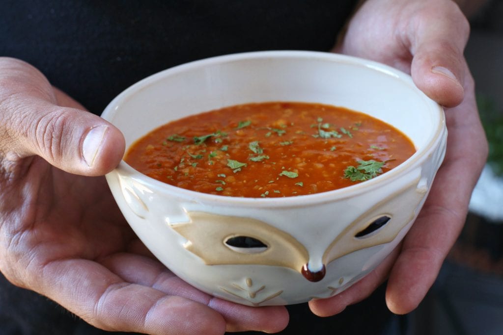Two hands hold a bowl of Ezo Gelin Corba is a spicy Turkish lentil soup made with red pepper paste an, bulgur wheat, and red lentils.