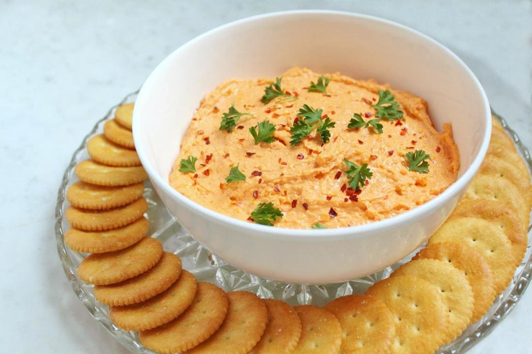 A white bowl containing orange Pimento Cheese dip surrounded by ritz crackers.