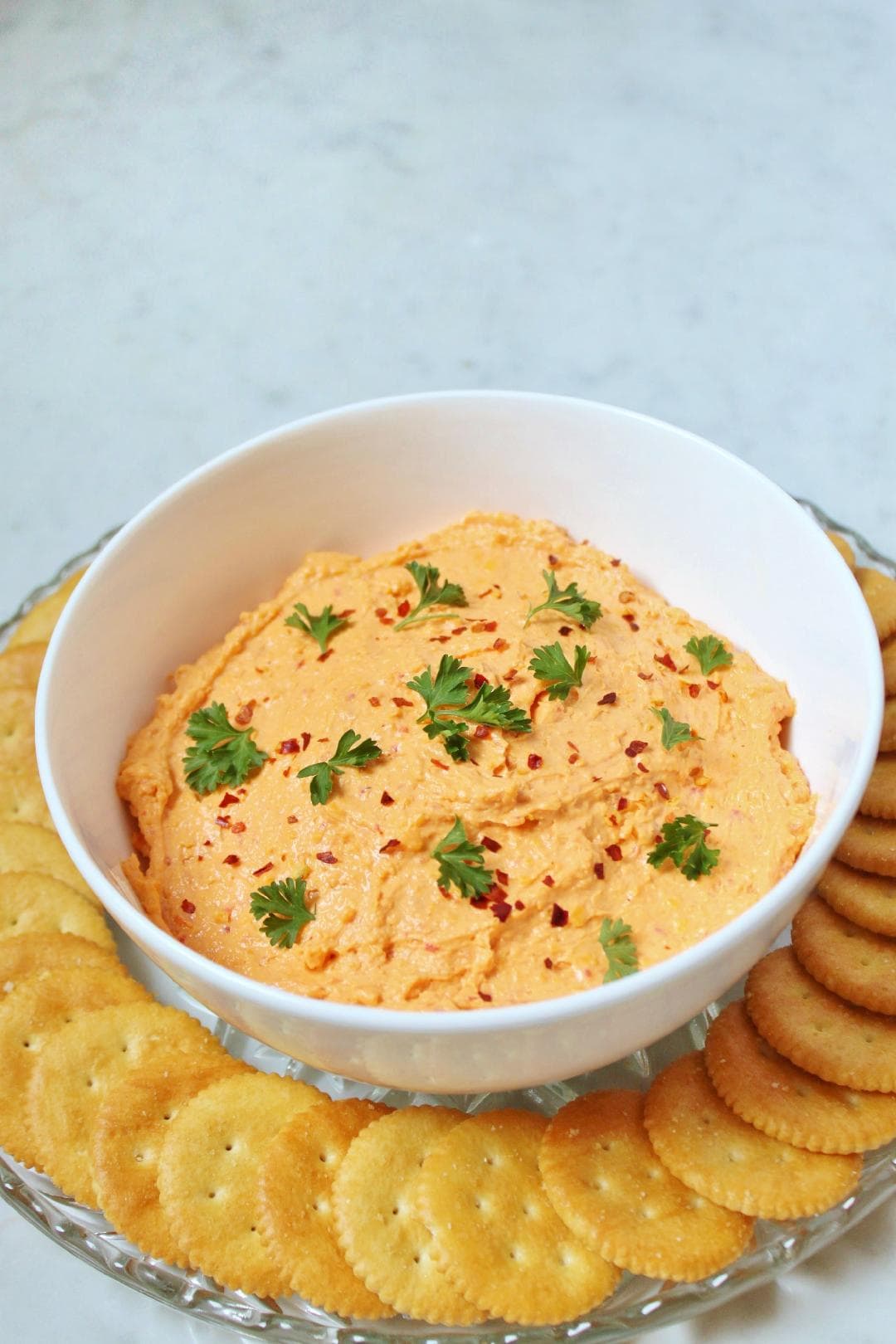 A white bowl containing orange Pimento Cheese dip surrounded by ritz crackers. 