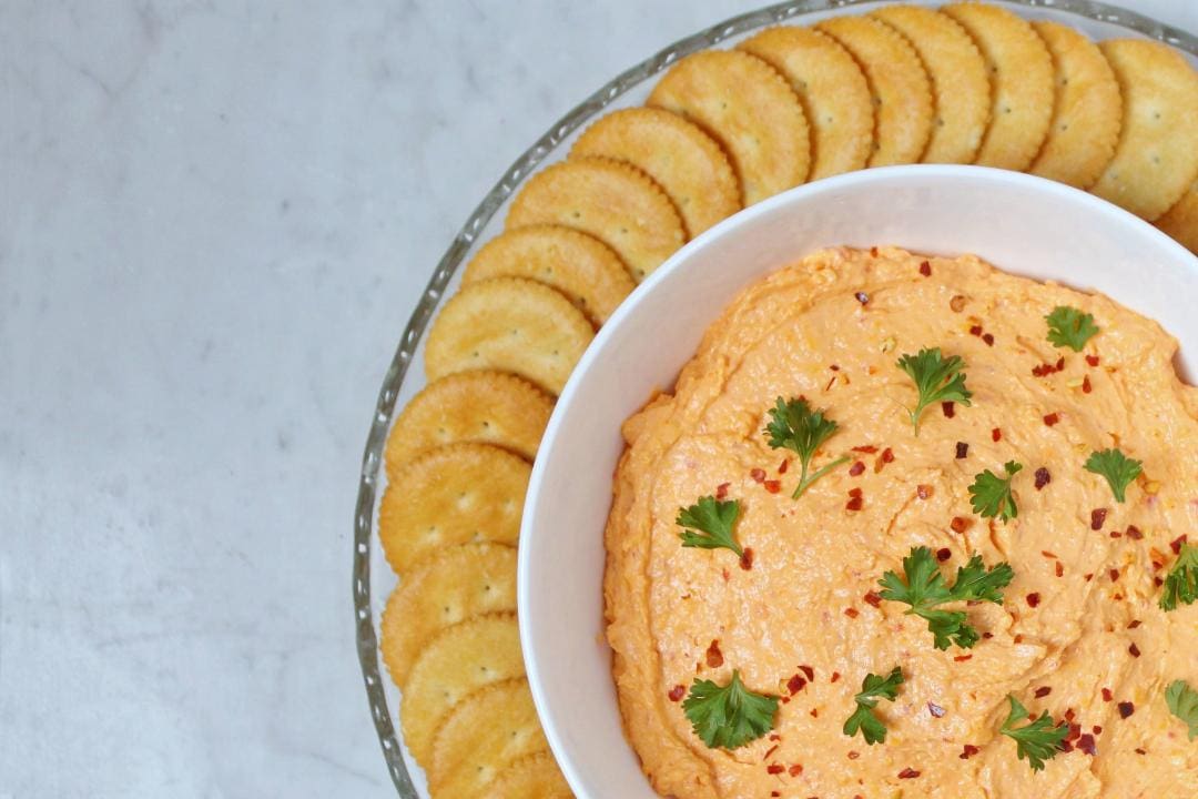 A white bowl containing orange Pimento Cheese dip surrounded by ritz crackers. 