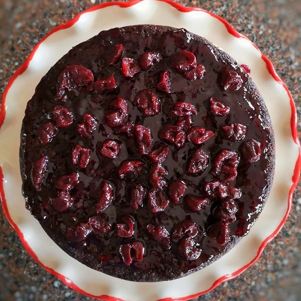 Overhead view of a cherry covered chocolate cake layer on a white plate with wavy red edges.