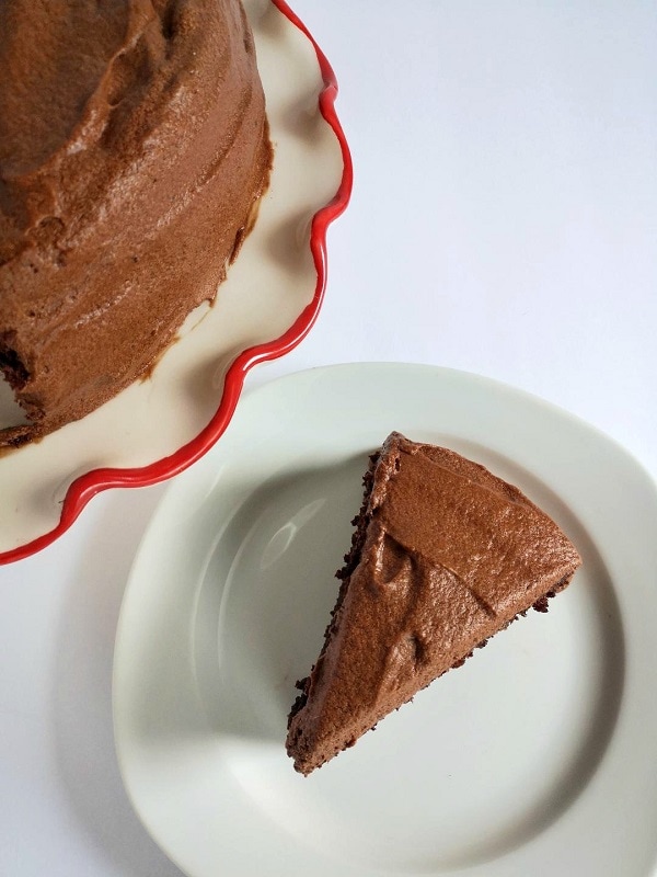 Slice of 2 layer chocolate cake on a white plate with a whole chocolate cake in the background