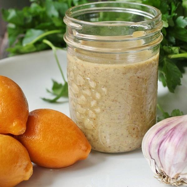 A vertical image of a jar of tahini salad dressing, preserved lemons, parsley, and garlic. 