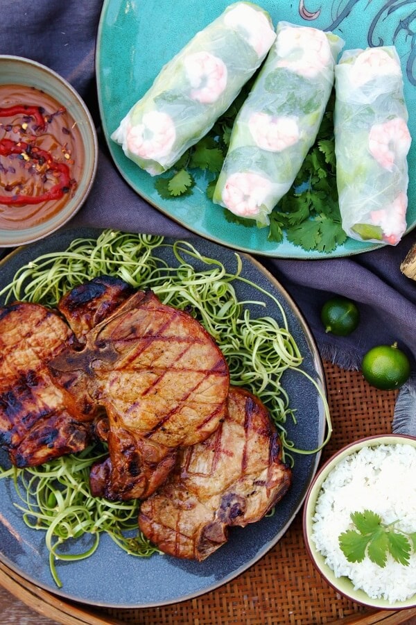 Three grilled Pork chops on a blue plate surrounded by a bowl of rice, spicy peanut sauce and salad rolls.
