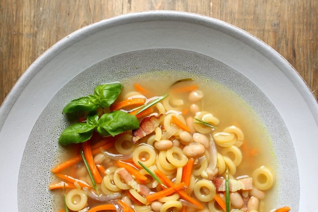 Noodle soup with beans, carrots, and bacon in a white bowl on a wooden background.