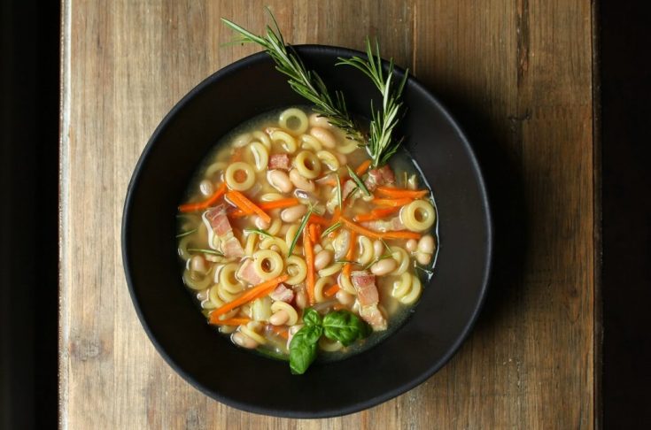 Noodle soup with beans, carrots, and bacon in a black bowl on a wooden background.