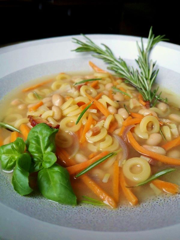 Pasta e Fagioli Noodle soup with beans, carrots, and bacon in a white bowl on a wooden background.