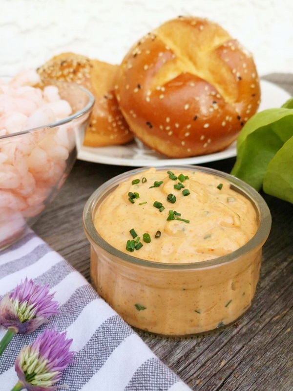 A small clear dish containing orange Remoulade sauce garnished with fresh chives. Surrounded by shrimp, fresh buns, and lettuce.