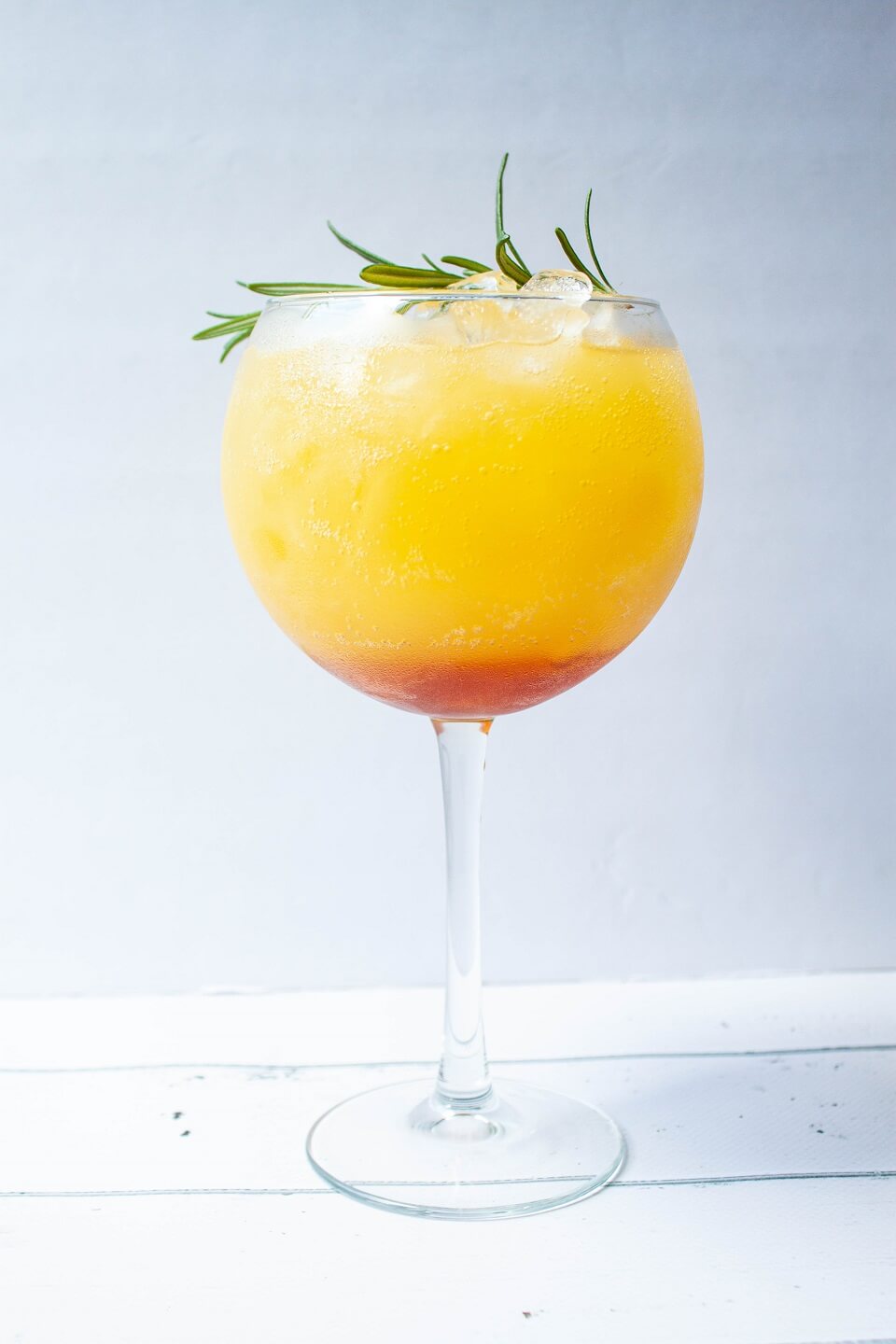 A single large wineglass containing an icy orange cocktail garnished with green rosemary.