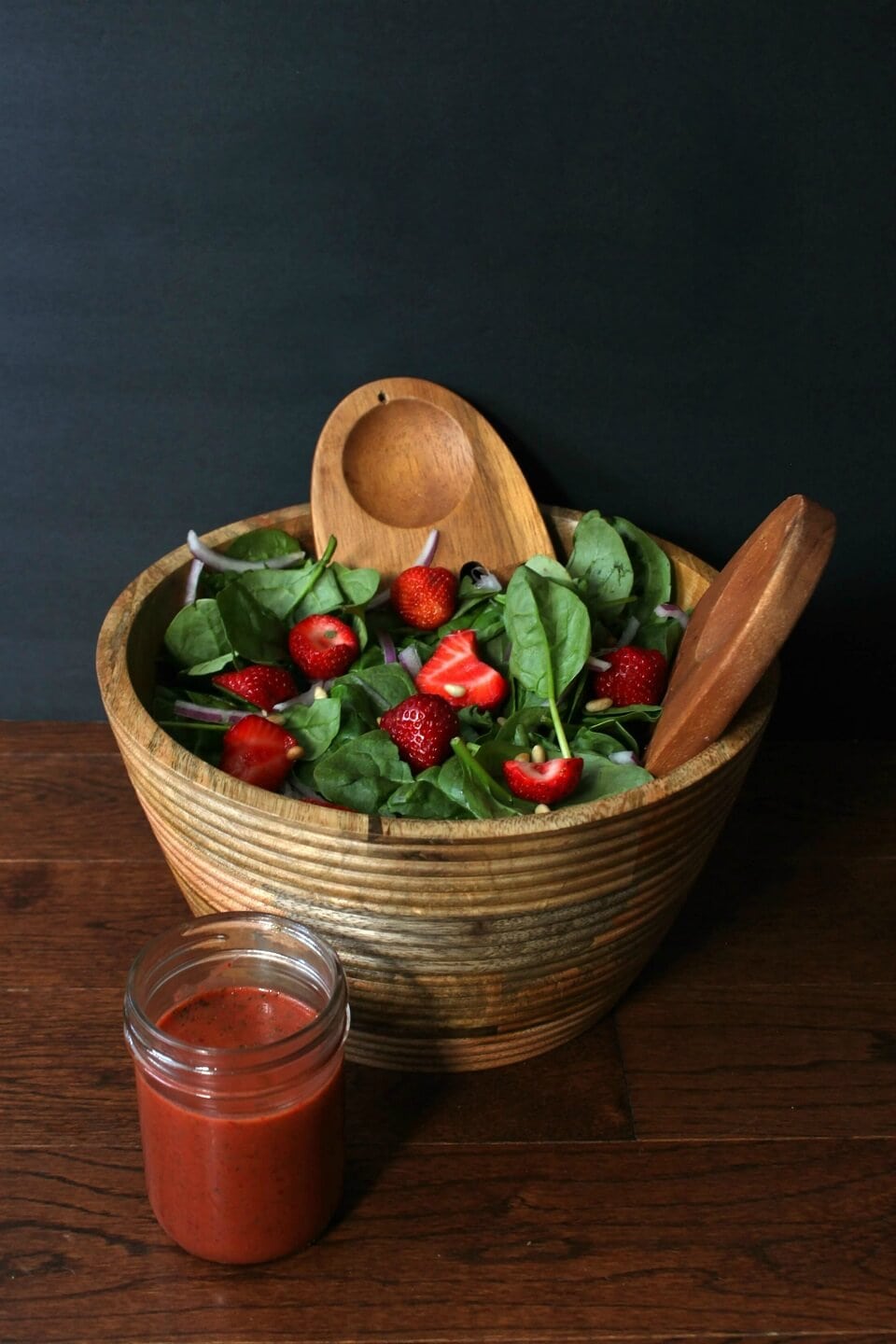 A wooden bowl containing green baby spinach, red strawberries, pine nuts and red onions.