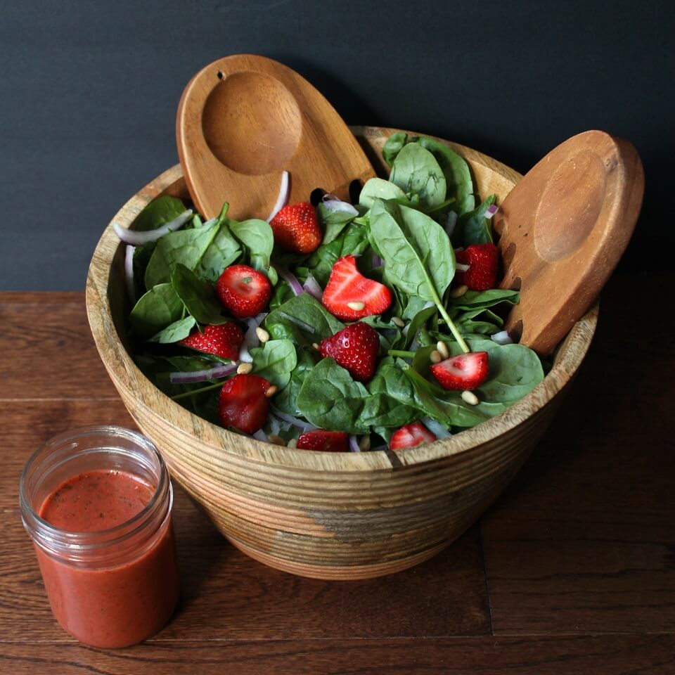 A wooden bowl containing green baby spinach, red strawberries, pine nuts and red onions.