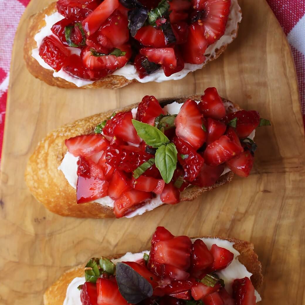 A wooden board holding Strawberry Basil Crostini: three slices of baguette topped with red strawberries and green basil.