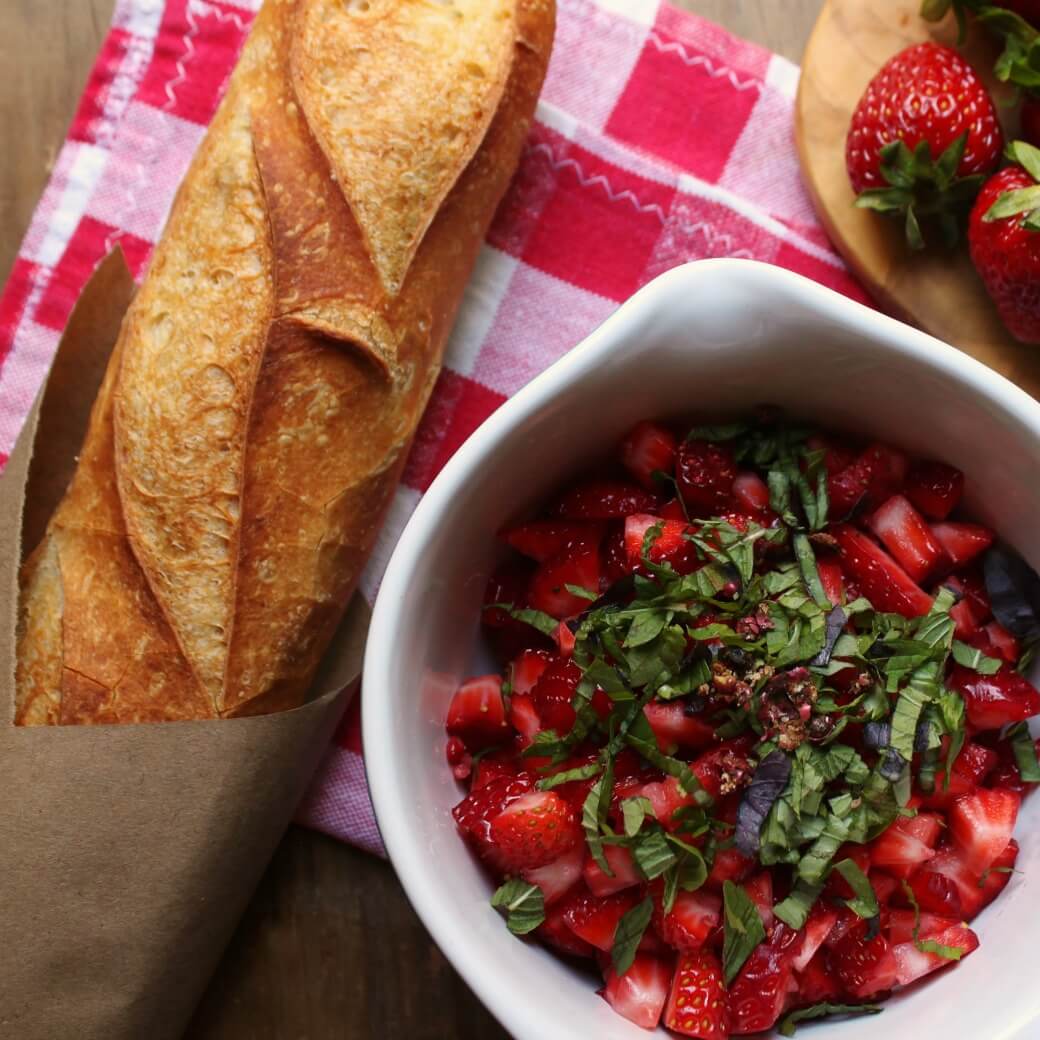 A bowl of chopped strawberries and basil beside a baguette and whole strawberries.