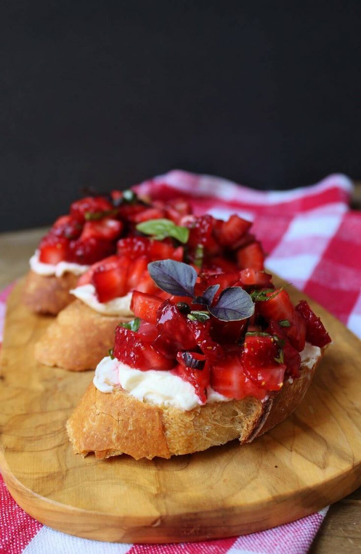 A wooden board holding three slices of baguette topped with red strawberries and green basil.