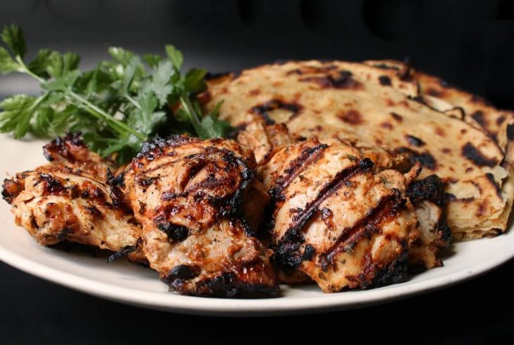 a white platter containing grilled chicken, paratha bread, and fresh cilantro.