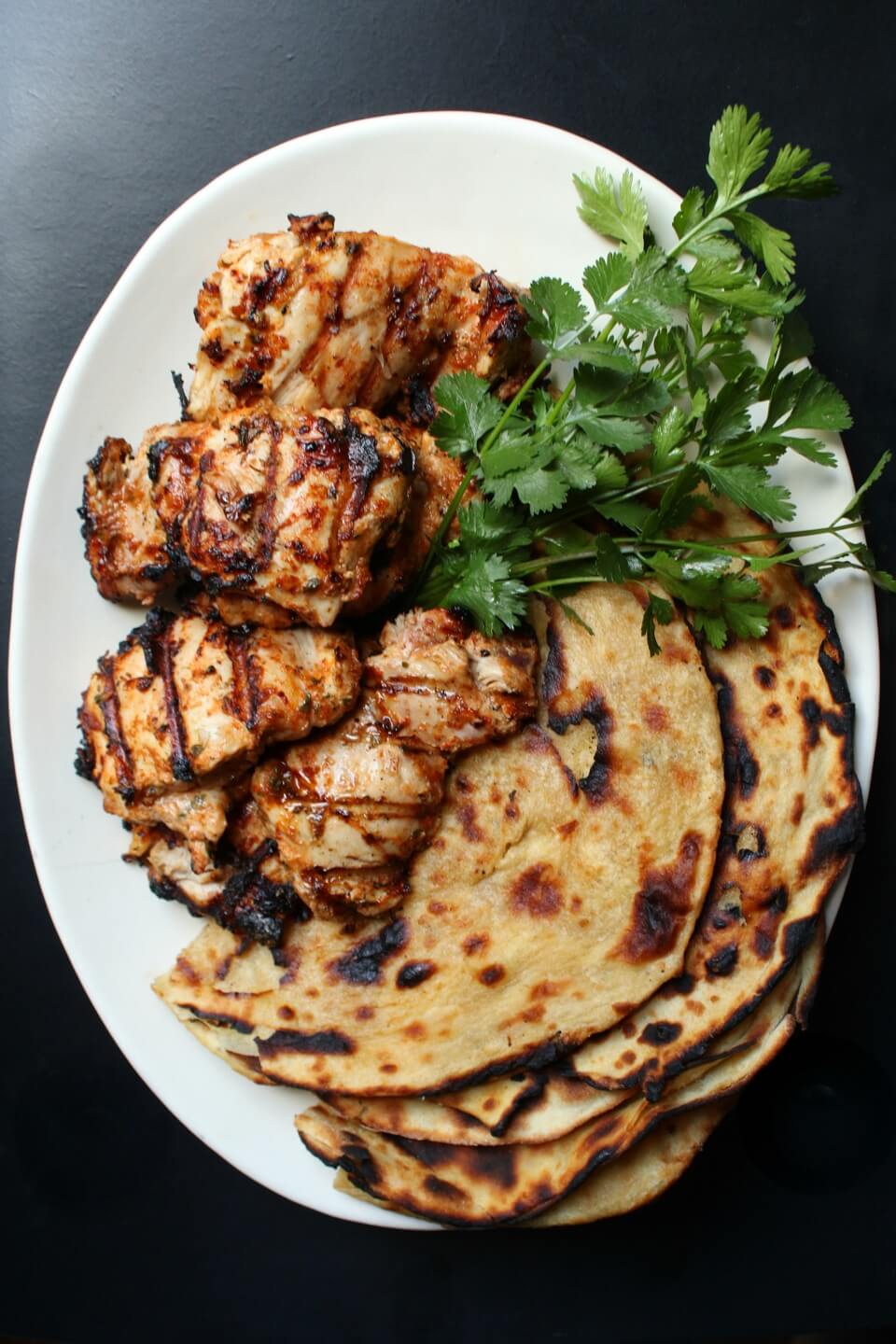a white platter containing grilled chicken, paratha bread, and fresh cilantro.