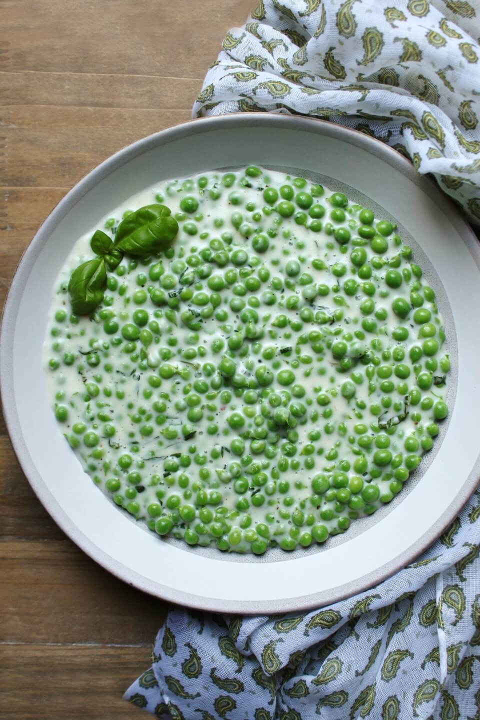 A bowl of green peas in a creamy sauce sits on a wooden table surrounded by a napkin with a green paisley print.