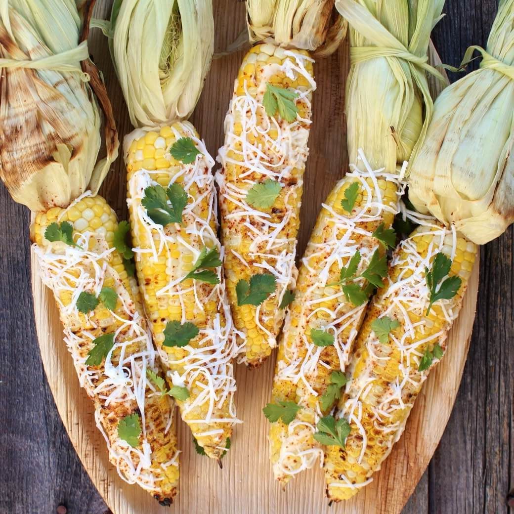 A wooden platter holding 5 cobs of Mexican Street Corn covered with mayonnaise and sprinkled with cheese, spices, and cilantro.