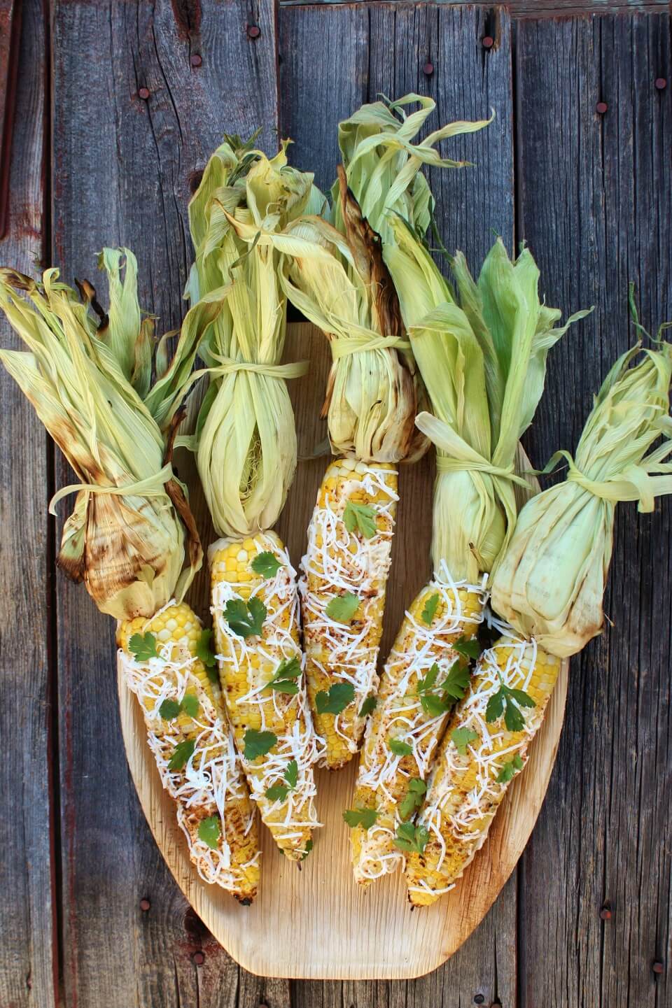 A wooden platter holding 5 yellow cobs of corn covered with mayonnaise and sprinkled with cheese, spices, and cilantro.