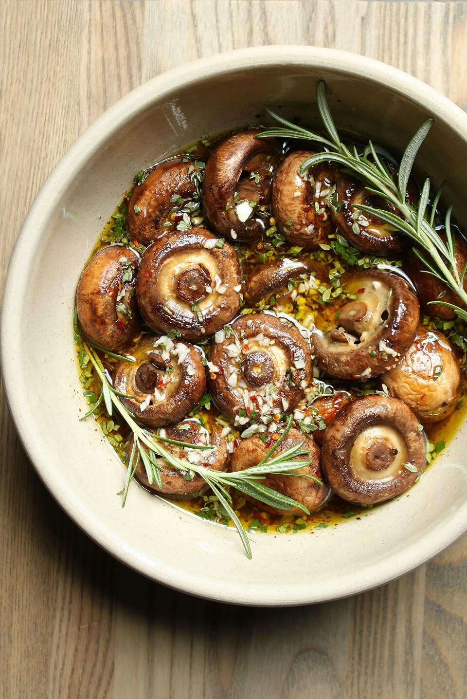 A crockery bowl full A crockery bowl full of brown marinated mushrooms and fresh rosemary sitting in a garlic and herb marinade.