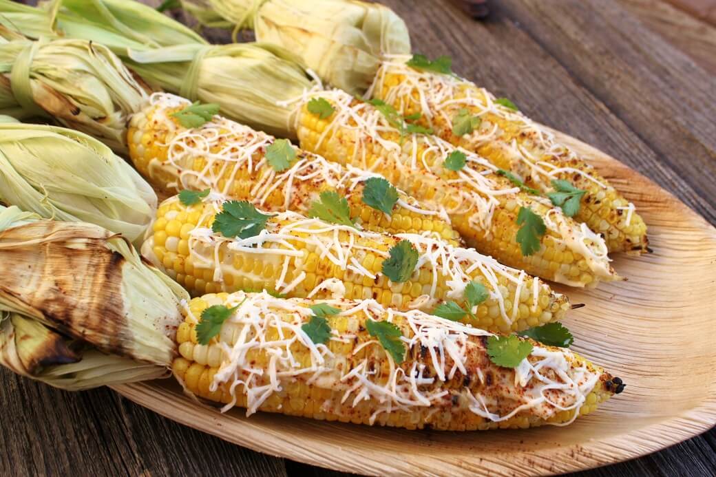 A wooden platter holding 5 yellow cobs of corn covered with mayonnaise and sprinkled with cheese, spices, and cilantro.