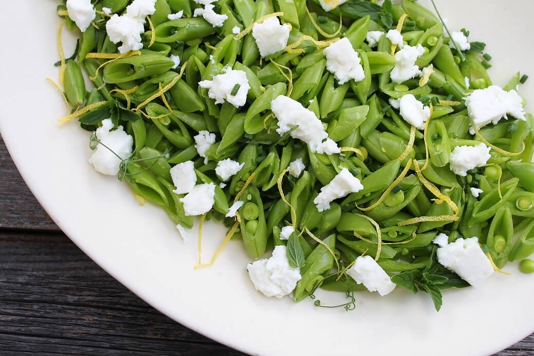 A white platter containing a bright green chopped snap pea salad, lemon zest, white feta, and herbs.