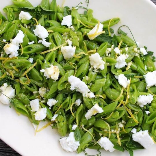 A white platter containing a bright green chopped snap pea salad, lemon zest, white feta, and herbs.