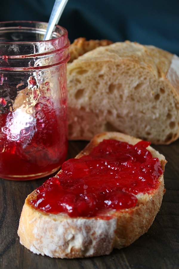 A slice of sourdough bread spread with ruby red sour cherry jam. Jar of jam and loaf of bread in the background.