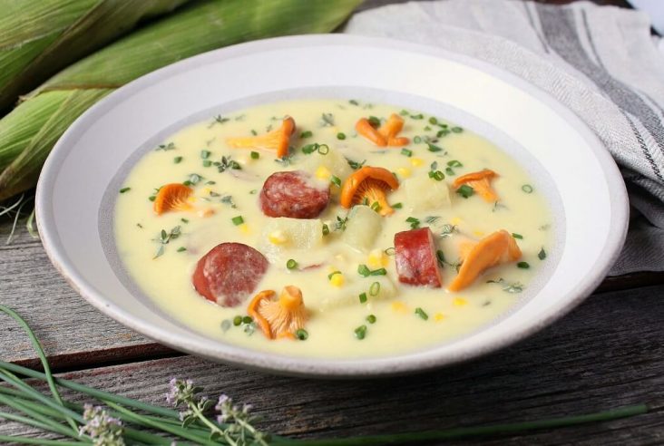 Hearty Farmer's Sausage Sweet Corn Chowder - A white bowl filled with creamy yellow soup with corn, sausage, potatoes and golden mushrooms garnished with fresh chives.