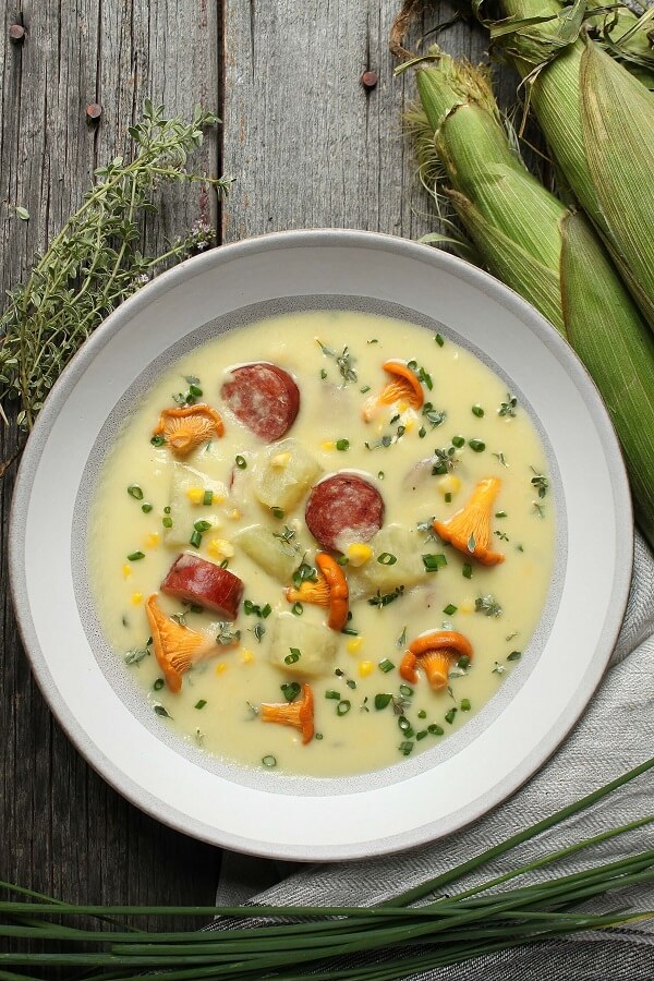 Hearty Farmer's Sausage Sweet Corn Chowder - A white bowl filled with creamy yellow soup with corn, sausage, potatoes and golden mushrooms garnished with fresh chives.