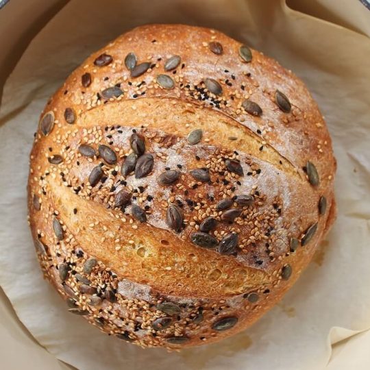 Pumpkin No Knead Bread -A boule of bread topped with pumpkin seeds in an ivory Dutch oven.