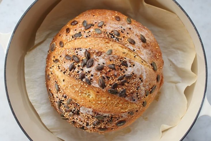 Pumpkin No Knead Bread -A boule of bread topped with pumpkin seeds in an ivory Dutch oven.