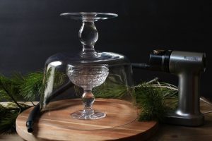 Cocktail smoking set up featuring a crystal coupe glass under a smoking cloche and a Breville smoking gun.