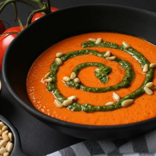 A black bowl of bright orange creamy tomato soup with a swirl of green pesto and scattered pine nuts.